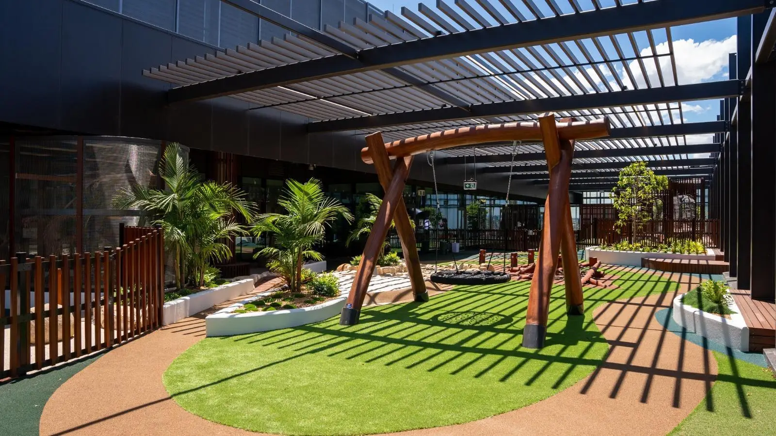 IMG-20260310-WA0008 - Michael Mitchell Greenspaces Outdoor playground in a modern building courtyard with a circular green turf patch and a large wooden A-frame swing under a metal pergola, surrounded by palm plants and planters.