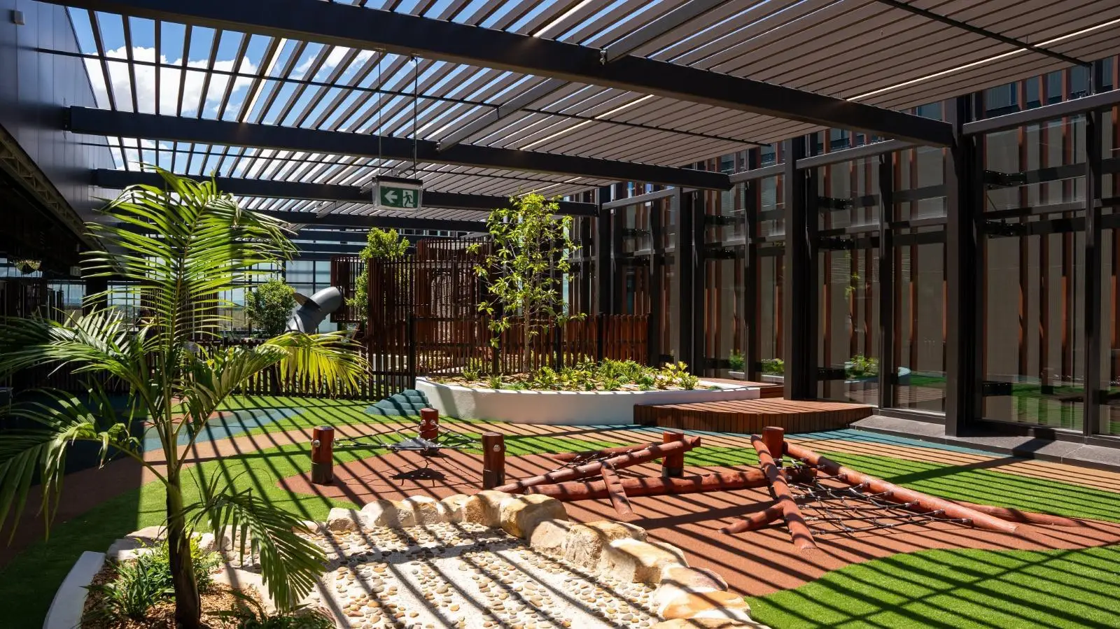 IMG-20260310-WA0010 - Michael Mitchell Greenspaces Sunlit modern courtyard with a translucent slatted roof, potted plants, and a wooden rope-play area on artificial turf nearby glass-walled buildings
