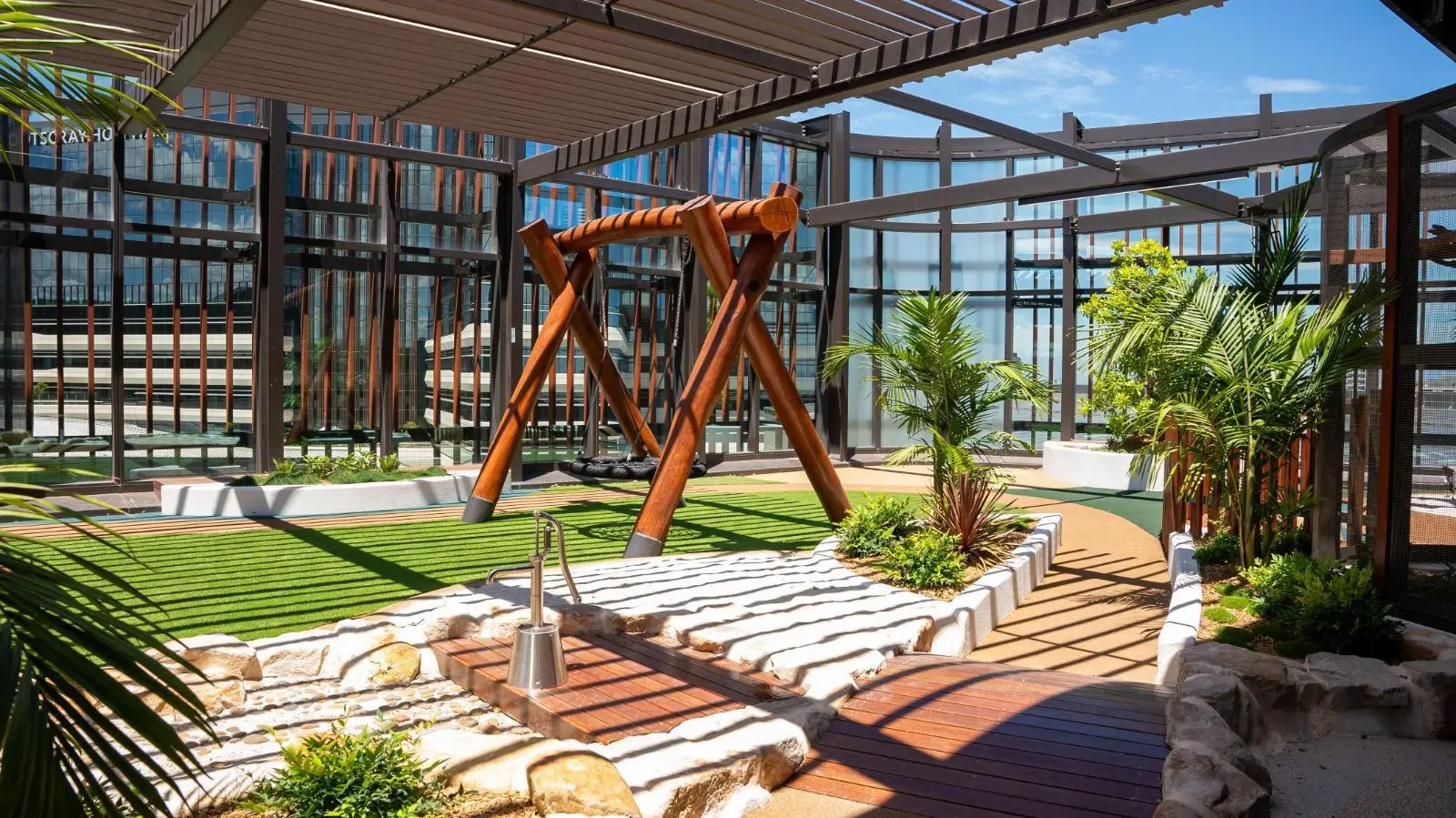 IMG-20260310-WA0017 - Michael Mitchell Greenspaces Rooftop garden with a wooden A-frame swing set, lush plants, and glass walls under a metal pergola for relaxation and recreation .