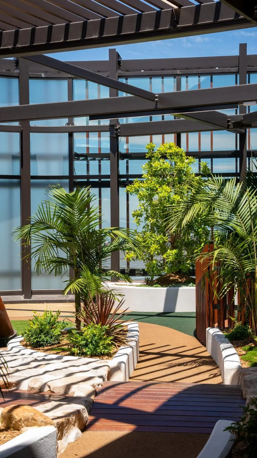 IMG-20260310-WA0020 - Michael Mitchell Greenspaces Indoor courtyard atrium with dark steel framework, glass walls, and a curved walkway flanked by tropical plants and planters
