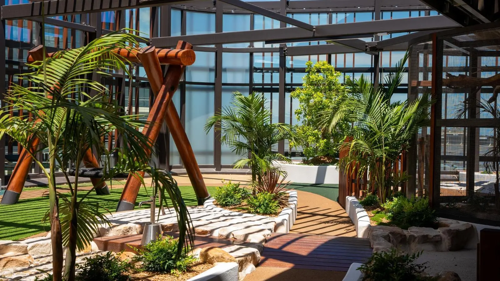 IMG-20260310-WA0022 - Michael Mitchell Greenspaces Sunlit courtyard with tropical plants, curved wooden walkway, and dark metal-framed glass walls.