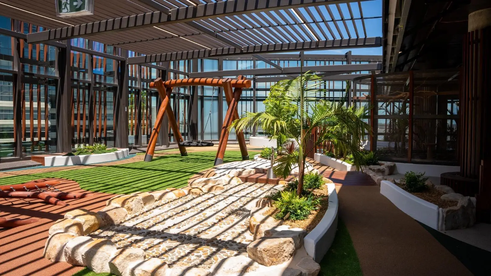 IMG-20260310-WA0023 - Michael Mitchell Greenspaces Rooftop courtyard featuring a wooden A-frame swing, stone pebble path, and palm plants under a striped pergola with glass walls nearby.