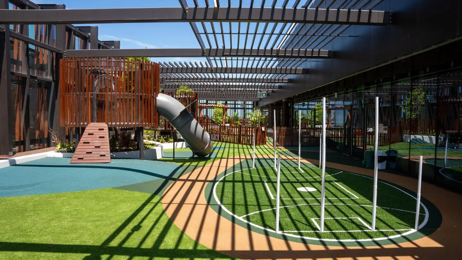 IMG-20260310-WA0036 1 - Michael Mitchell Greenspaces Rooftop playground with a curved metal slide, wooden climbing ramp, and a circular artificial turf area with goal lines, shaded by a slatted metal pergola.