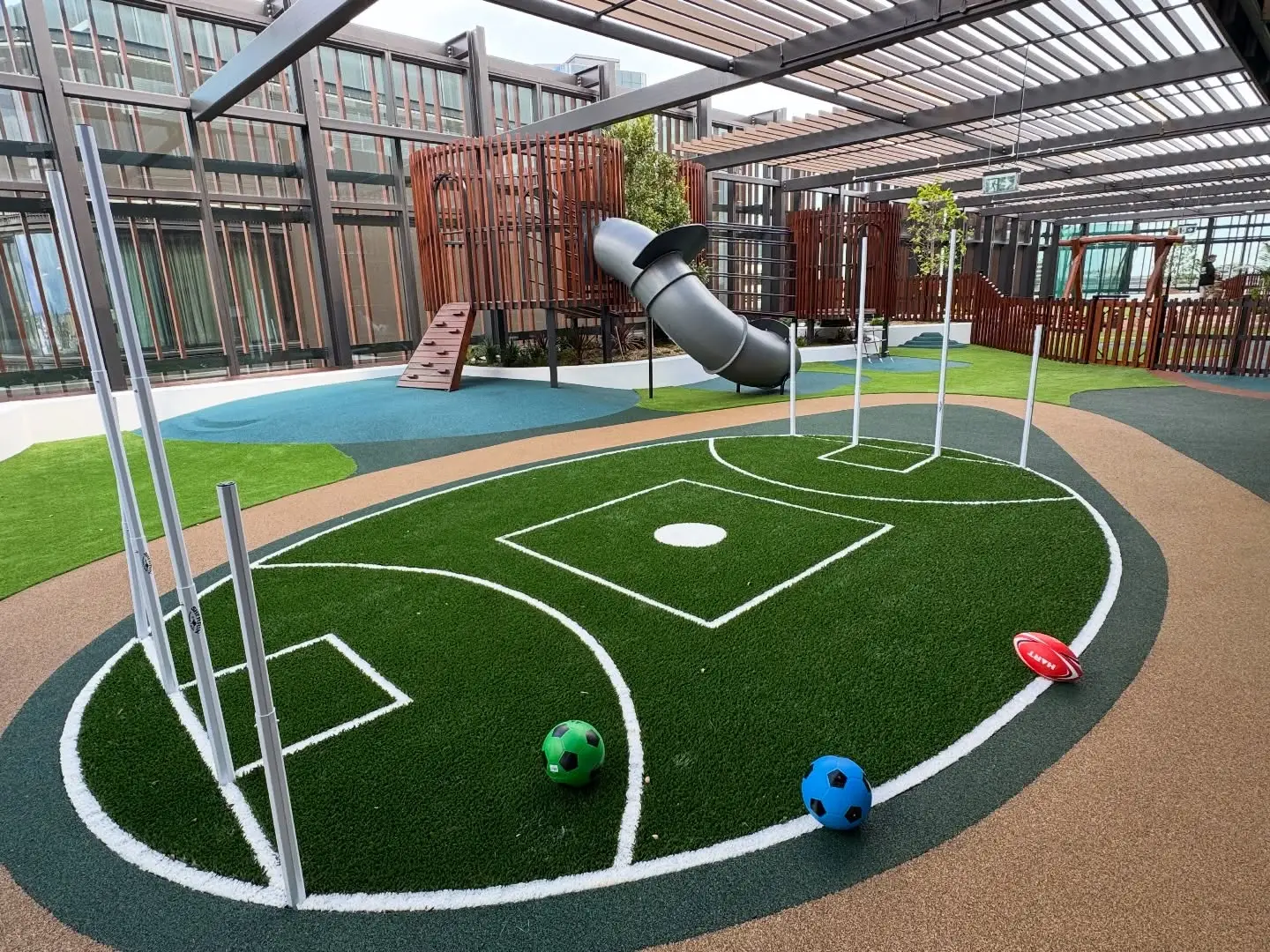 Kool Beanz Bulldogs - Michael Mitchell Greenspaces Rooftop playground with a synthetic baseball field, curved slide, and climbing wall under a glass roof edge. Three colored balls sit on the turf.