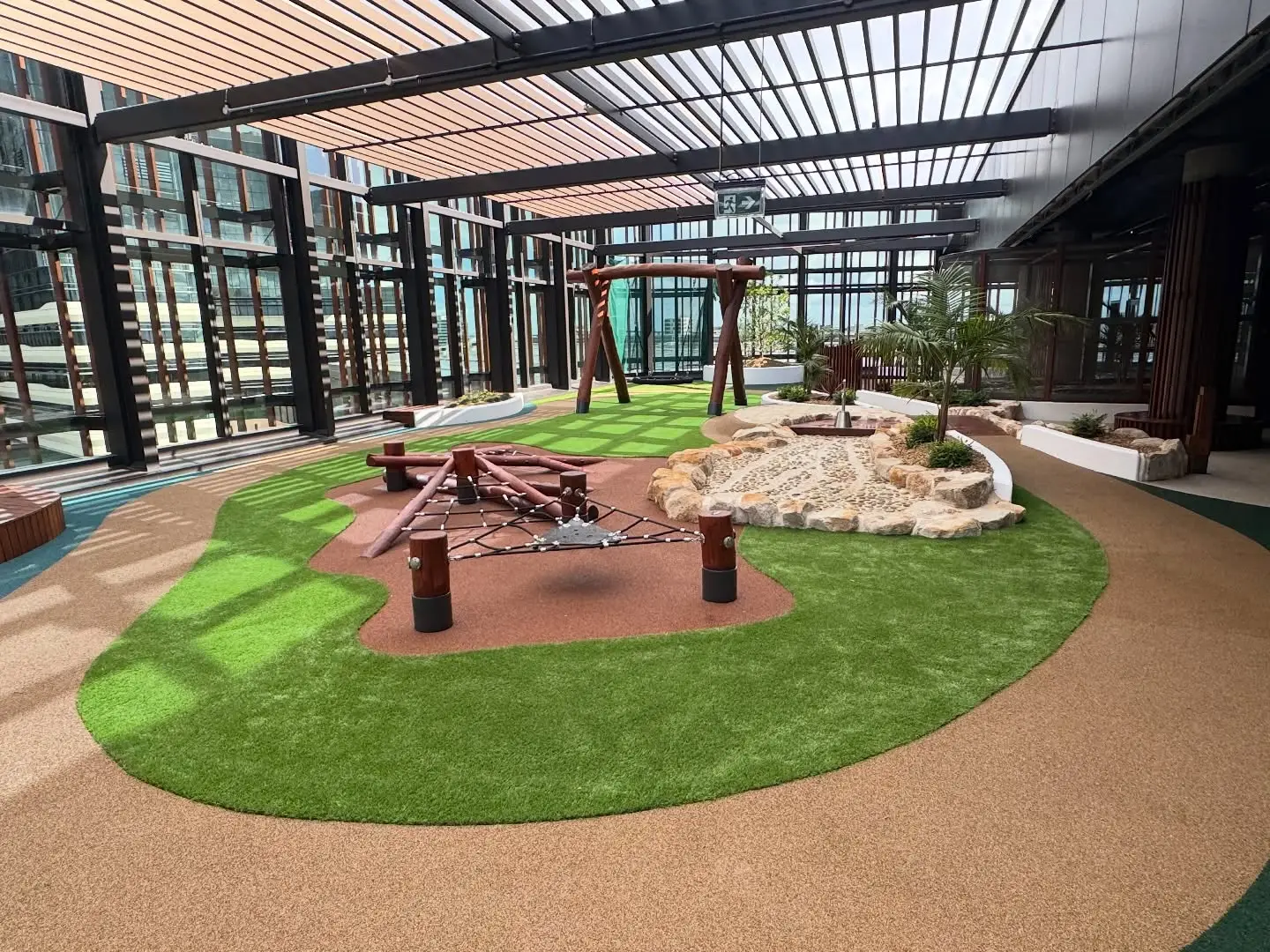 Aarons Outdoor_Kool Beanz Bulldogs 5 - Michael Mitchell Greenspaces Indoor glass-walled courtyard playground with green artificial turf, brown rubber paths, wooden climbing frame and rock landscaping.