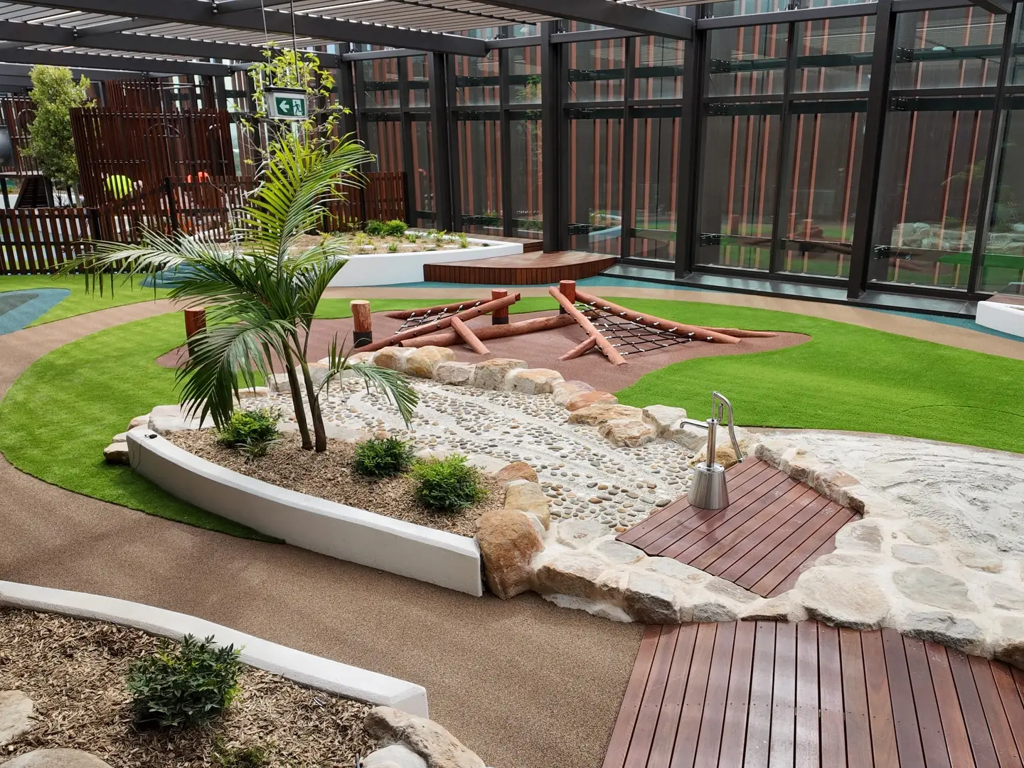 - Michael Mitchell Greenspaces Indoor courtyard playground with artificial grass, a rock garden, and a wooden climbing structure under a glass ceiling.